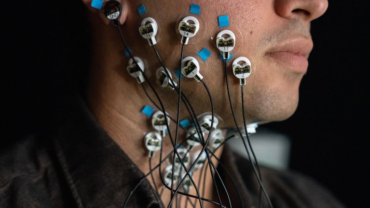 Close-up of a participant’s face and neck with multiple electrodes attached using blue medical tape. The electrodes are connected to thin black wires, leading to a monitoring device, highlighting a scientific study on muscle or nerve activity